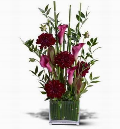 Burgundy calla blossoms arranged with winecolored dahlias in a rectangular glass vase lined with equisetum stalks in grass.