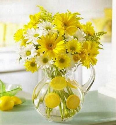 Bouquet of fresh yellow gerbera daisies, yellow and white daisy poms, solidago and more in a clear acrylic pitcher.