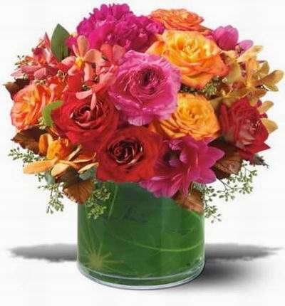 Mix of dahlias peonies roses and orchids in a range of pinks oranges and reds in a clear leaflined cylinder.